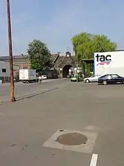 La photographie montre une plaque d'égout ronde entourée d'un carré en béton et comportant une petite plaque métallique de dix centimètres de côté approximativement, il s'agit du puits Nord de la fosse du Pavé à Anzin. L'environnement est constitué d'un parking relativement vide.