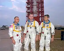 Trois hommes en combinaison spatiale, sans casque, devant une tour de lancement.