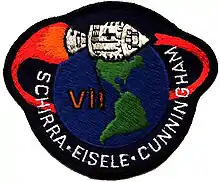 Écusson représentant un vaisseau spatial volant au-dessus du continent américain. Un gros chiffre romain VII et le nom des trois astronautes.