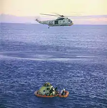 Un hélicoptère survole un vaisseau spatial et un canot pneumatique flottant sur la mer.