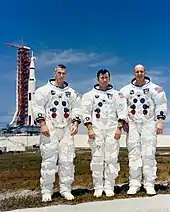 Trois hommes debouts en combinaison spatiale. Fusée sur site de lancement à l'arrière plan.