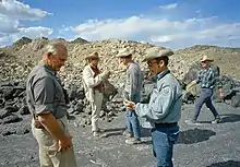 Photographie en couleur avec, au premier plan, John Young discutant avec William R. Muehlberger, au second plan, Charles Duke discutant avec David Wones. Anthony W. England est visible sur la droite de la photo. Des monticules de roches sont visibles à l'arrière-plan.