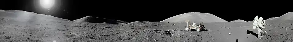 Panorama de la surface lunaire. Un rover et un astronaute sont visibles. Le soleil est en haut à gauche de l'image.