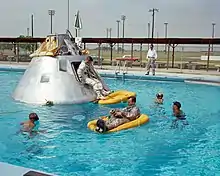 Photographie en couleur d'hommes en combinaison s'entraînant à s'extraire d'une capsule flottant dans une piscine.