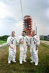 Trois hommes en combinaison spatiale, sans casque, devant une tour de lancement.