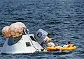 Photographie en couleur de la capsule d'Apollo 7 flottant sur la mer et les astronautes en sortant pour rejoindre un bateau pneumatique jaune.