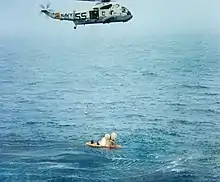 Un hélicoptère volant au-dessus d'un vaisseau spatial et d'un canot pneumatique, dans la mer.