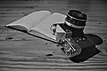 Photographie en noir et blanc. Un appareil photo Zenit-E repose pour partie, objectif pointant vers le haut, sur un livre ouvert. Le livre, illisible, repose à-plat sur un support ayant l’aspect du bois.