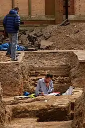 Photo d'une tranchée prise à hauteur du sol. Un homme se trouve au fond de la tranchée et examine des documents, tandis que deux personnes l'observent depuis le bord de la tranchée.