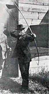 Photo d'un homme en costume et haut de forme s'apprêtant à décocher une flèche avec un arc.