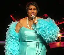 A woman stands on stage and sings into a microphone. She wears a blue dress and has a blue feather boa around her arms.