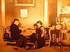 Un homme et une femme assis sur un canapé en conversation dans un salon richement meublé. L'homme avec des guêtres lit un manuscrit.