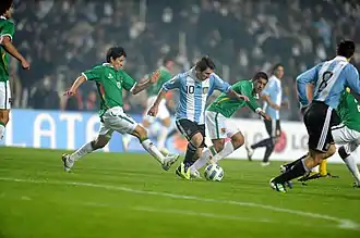 Photo d'un match de football, un joueur en bleu et blanc court avec le ballon poursuivi par deux joueurs en vert