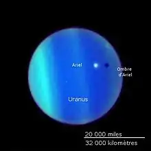 La planète Uranus vue par le télescope Hubble, son atmosphère forme des bandes bleues électrique et vertes. Ariel apparaît comme un point blanc flottant au-dessus et jette un voile sombre au-dessous.