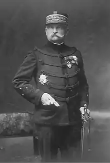 Photographie monochrome d'un homme moustachu, debout, de face, portant une tenue de général, le képi et l'épée au côté.