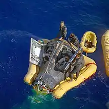 Photographie en couleur d'Armstrong et Scott dans leur capsule attendant avec l'équipe de sauvetage l'arrivée de leur navire.