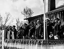 Hommes en costumes et uniformes debout sur une estrade décorée en train de saluer.