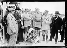 Photographie en noir et blanc de deux coureurs entourés par un gendarme et des officiels à l'arrivée d'une course, l'un tenant un bouquet de fleurs à la main.