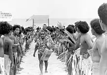 Homme vêtu d'une chemise et d'un short marchant sur la plage entre deux rangées d'hommes en pagne