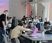 Photographie en couleurs d'une vaste pièce lumineuse et remplie de tables dans laquelle plusieurs personnes se déplacent d'un groupe de travail à un autre.