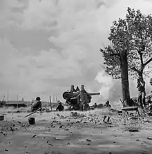 Photographie en noir et blanc montrant des soldats sur une plage autour d'un canon qui tire ; une volute de fumée s'en échappe. À droite, près d'un arbre, un soldat se bouche les oreilles face au bruit du tir.