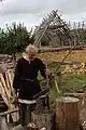 Photographie montrant un homme costumé, équipé d'un maillet et d'un départoir, se préparant à fendre une bûche, avec un bâtiment en pierres en construction, surmonté d'une charpente formée de troncs.