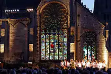 Sons et lumières, Folgoët 2010. Devant un public nombreux, groupe de 22 personnes. En arrière-plan, les vitraux de l'église, sembla,t éclairés de l'intérieur, occupent la quasi-totalité de la photo, en hauteur