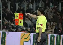 Photo d'un arbitre de touche en jaune levant son drapeau avec les tribunes en fond.