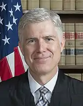 Portrait de Neil Gorsuch dans sa robe de juge devant une bibliothèque et le drapeau américain.