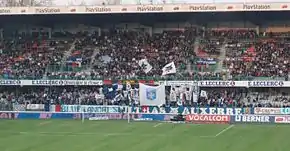 Photographie montrant la tribune d'un stade de football, remplie de supporters agitant des drapeaux