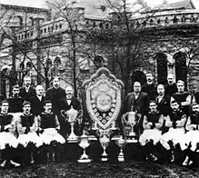 Photo en noir et blanc d'une équipe de football posant autour de son blason et de ses trophées au centre.