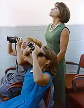 Deux filles regardent vers le ciel avec des jumelles, une femme derrière elles regarde dans la même direction.