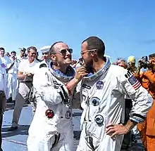 Deux hommes en combinaison spatiale se félicitent. Foule en arrière-plan.