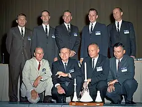 Photographie en couleur du groupe groupe d'astronautes 2 : les hommes en costume sont alignés sur deux rangées avec au premier plan des maquettes de vaisseaux spatiaux.