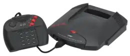 Photo de la console Jaguar à laquelle est branchée une manette de jeu, composée d'une croix directionnelle, deux boutons centraux et trois gros boutons rouges à droite, et de douze boutons dans sa partie centrale