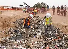 Photographie de deux ouvriers excavant des déchets de la décharge devant des spectateurs.