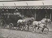 Photo en noir et blanc représentant une voiture tirée par quatre chevaux gris.