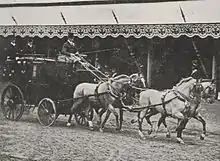 Photo d'une voiture sur laquelle sont assises trois personnes attelée à quatre chevaux.