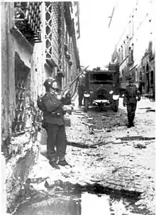Image en noir et blanc avec un soldat pointant son fusil vers le haut, dans une rue endommagée, avec en arrière-plan un autre soldat et un camion d'armée