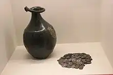 Présentation d'une cruche et d'un tas de pièces de monnaies dans une vitrine.