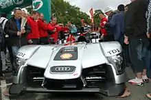 Photo d'une voiture de sport-prototype, grise et noir. Une foule, des arbres et un ciel gris en arrière plan.