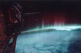 Des faisceaux colorés s'échappent de l'atmosphère terrestre, à droite de l'image ; à gauche, la soute de la navette spatiale est ouverte.
