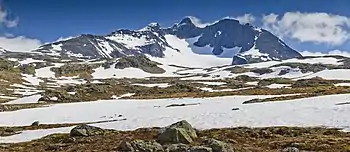 Paysage plat avec névés au premier plan et montagne en arrière-plan avec un glacier entouré de parois rocheuses verticales.