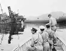 Vue en noir et blanc de l'avant d'une barque avec 4 soldats. En arrière-plan un navire naufragé.