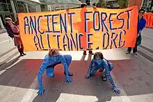 Deux personnes entièrement peintes en bleues sont accroupis devant un panneau orange avec écrit Ancient Forest Alliance.