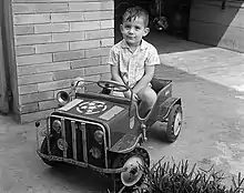 Photographie en noir et blanc d'Ayrton Senna à l'âge de trois ans sur une petite voiture pour enfant.