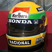 Photographie couleur du casque de Senna, majoritairement jaune, avec une bande verte et des sponsors, au musée Honda.
