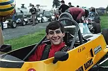 Photo en couleur d'un jeune homme brun, de face, souriant, habillé en rouge, posant sans son casque dans le baquet d'une Formule Ford jaune.