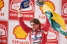 Photographie de Senna, vu de trois-quarts, en combinaison rouge, saluant la foule, les bras aux ciel, drapeau brésilien à la main, sur le podium.