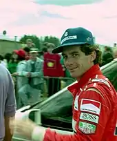 Photo d'Ayrton Senna en plan poitrine, corps de profil et tête de face, devant des spectateurs, passant dans les paddocks du circuit.